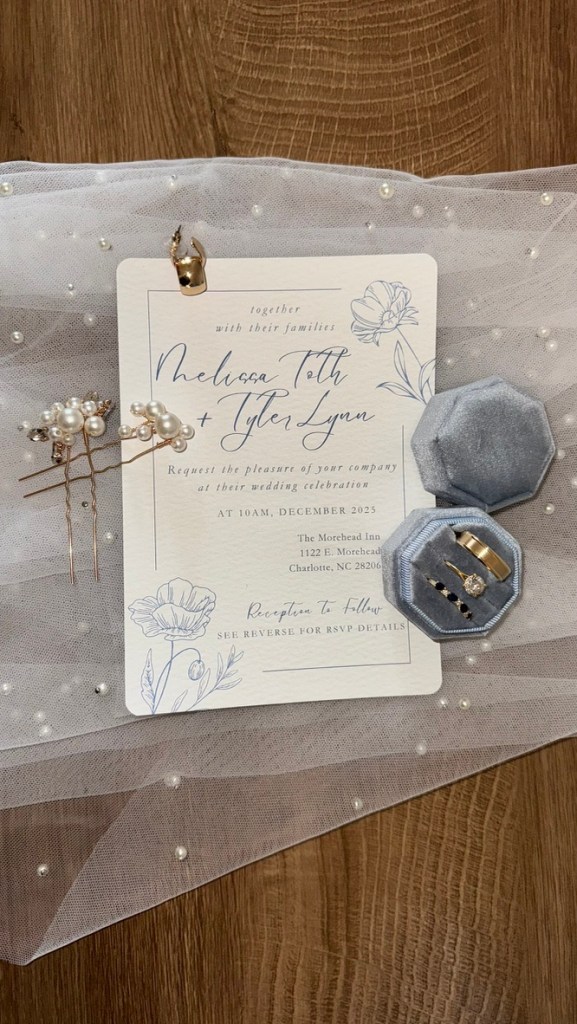 A wedding invitation card featuring the names Melissa Toth and Tyler Lynn, set on a soft tulle background adorned with pearls. Accompanying the invitation is a light blue hexagonal ring box containing wedding rings, alongside decorative hair pins.
