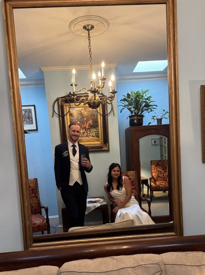 A couple dressed in formal attire, with the man in a suit and the woman in a wedding dress, posing in front of a large mirror. The setting features elegant furniture and decorative elements.