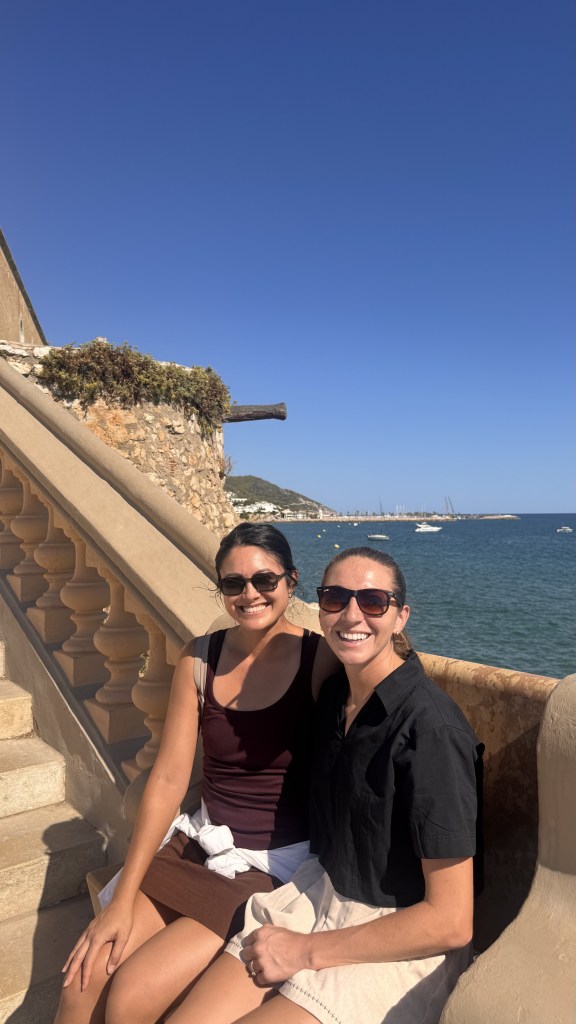 Two women sitting on stairs with a coastal view in the background, wearing sunglasses and smiling.