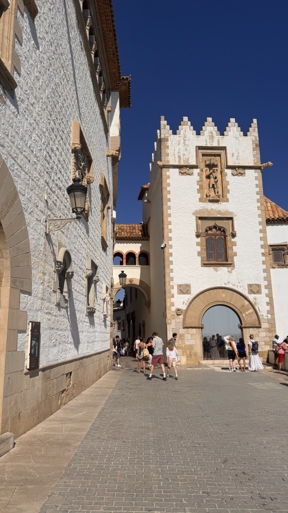 A historic street scene featuring a white stone building and a castle-like structure with decorative features, under a clear blue sky. Pedestrians are visible along the cobblestone path, adding to the lively atmosphere.