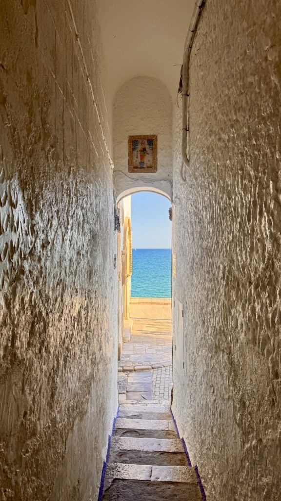 Narrow stone staircase leading through a whitewashed corridor to a view of the ocean and coastline.